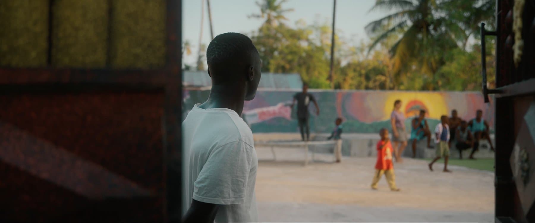 A young performer looking out over the training yard as children play near the mural wall