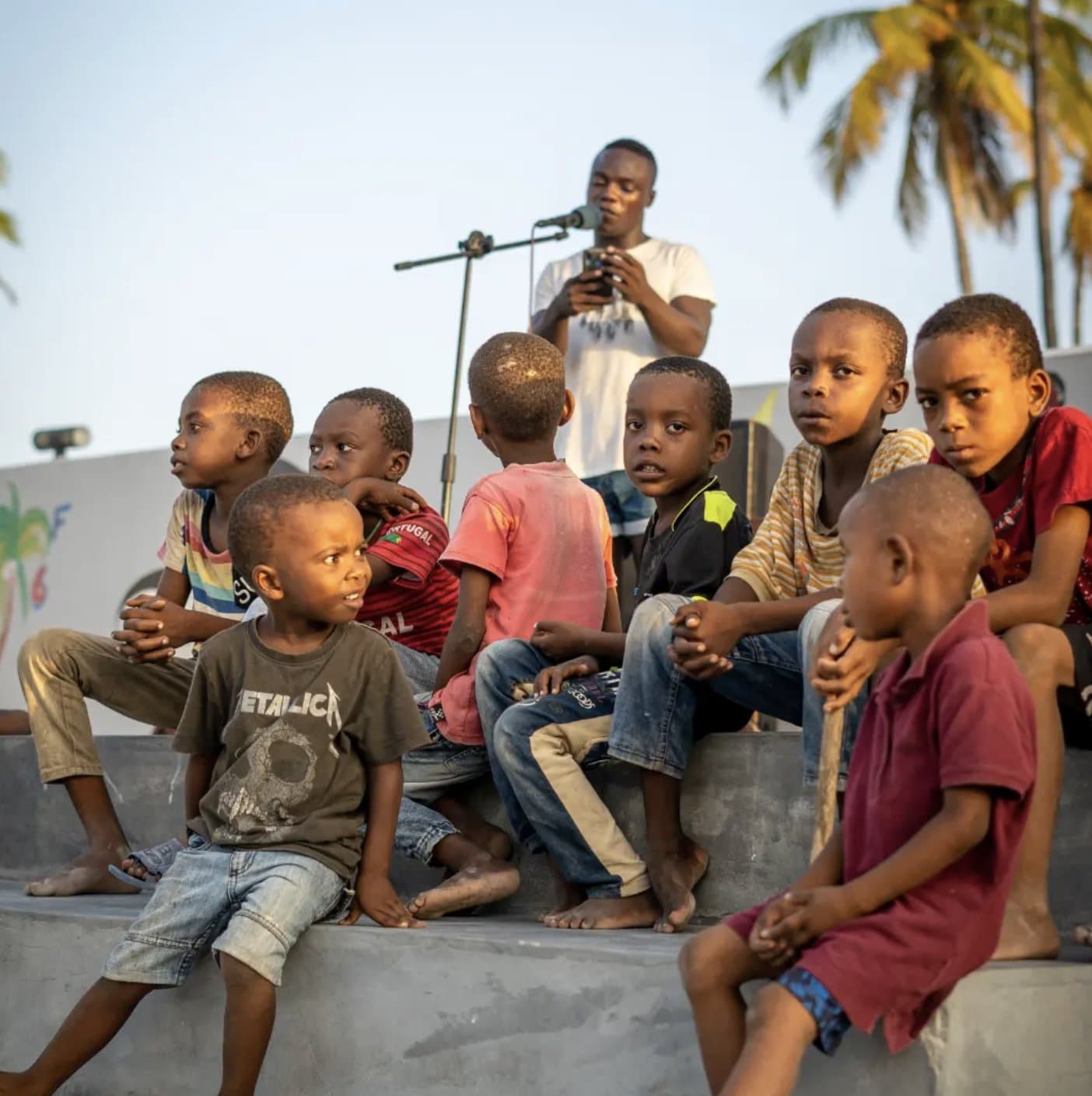 Children from the Move Zanzibar community sitting together, listening to a young performer with a microphone, palm trees behind them